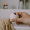 A hand holding a white bath bomb with dried flower petals on top, with a beige textured towel and a softly blurred lit candle in the background.