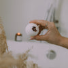 A hand holding a white bath bomb with dried flower pieces, set against a softly blurred bathroom background with a lit candle and neutral decor.