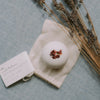 A white bath bomb with dried red flowers on top, resting on a small cream fabric pouch next to a printed card, styled with dried lavender on a light blue textured background.